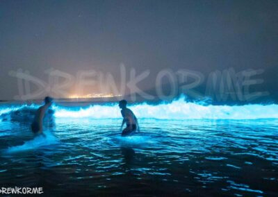 Dos personas bañándose en el mar de ardora en Area das Vacas, Cabana de Bergantiños, con olas bioluminiscentes iluminando el agua en la Costa da Morte, Galicia