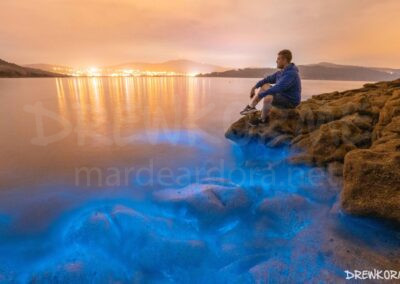 Persona sentada sobre una roca observando el mar de ardora en la playa de Balares (Ponteceso, Costa da Morte, Galicia)