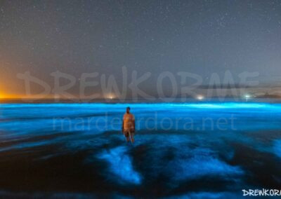Persona en el mar de ardora en la playa de Carnota con olas bioluminiscentes, cielo estrellado y el faro de Fisterra al fondo, Costa da Morte (Galicia)