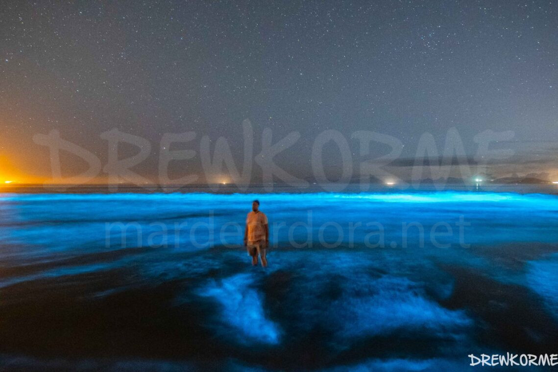 mar-de-ardora-carnota-costa-da-morte-galicia-drewkorme- Persona en el mar de ardora en la playa de Carnota con olas bioluminiscentes, cielo estrellado y el faro de Fisterra al fondo, Costa da Morte (Galicia)