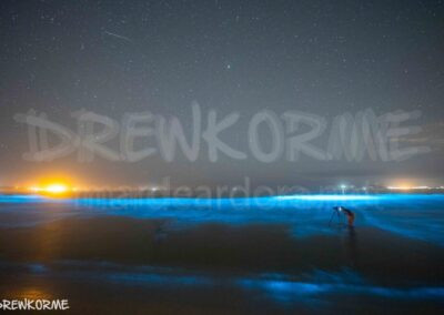 Fotógrafa captando el mar de ardora desde el agua en la playa de Carnota, Galicia, con bioluminiscencia azul iluminando el mar bajo el cielo estrellado