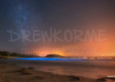 Mar de ardora y Vía Láctea en la playa de A Ermida (Corme, Costa da Morte, Galicia) con bañistas en el agua y bioluminiscencia en las olas