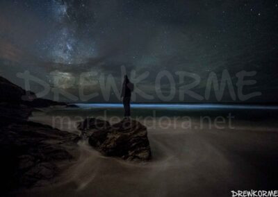 Mar de ardora y Vía Láctea en la playa de Mar de Fora en Fisterra, Costa da Morte (Galicia), con una persona sobre las rocas observando el cielo nocturno